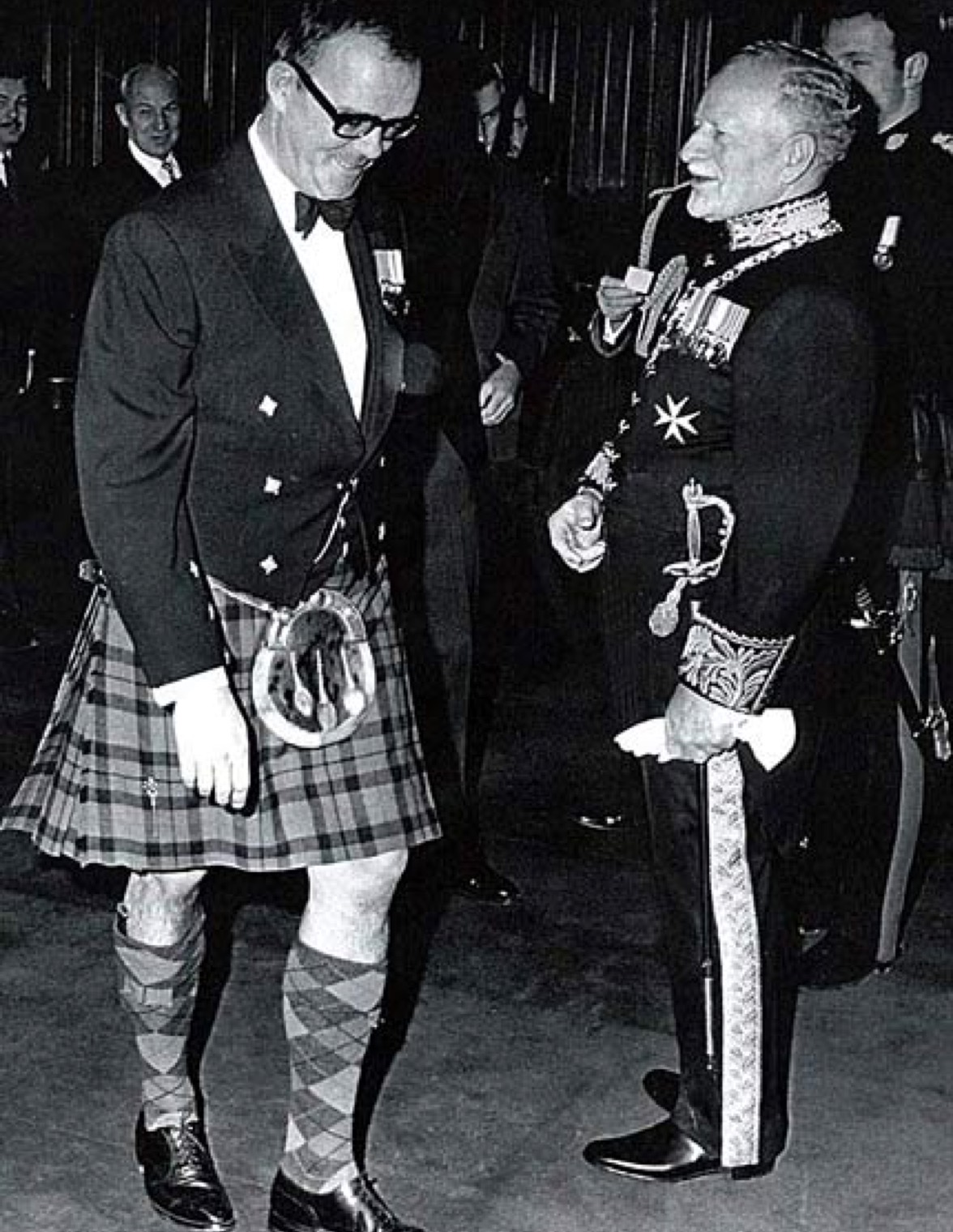 Deux hommes se trouvent au premier plan. L'homme de gauche porte un kilt, l'homme de droite porte un uniforme militaire.